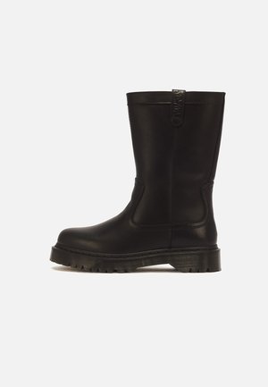 Schwarzer mittellanger Lederstiefel mit dicker profillierter Sohle und Zuglasche oben, von der Seite auf einfarbigem Hintergrund gezeigt.