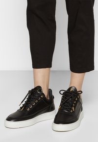 Černé kožené tenisky s bílou podrážkou, zlatými očky a texturovanými akcenty. Tkaničky jsou svázány v standardní konfiguraci.