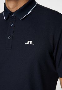 Navy pikétröja gjord av texturerad tyg med en kontrasterande vit krage och en liten vit logotyp på vänster bröst.