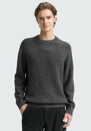 Junger Mann trägt einen dunkelgrauen gerippten Pullover und schwarze Hose, steht mit einer Hand in der Tasche vor einem einfarbigen hellen Hintergrund.