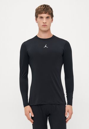 Jaunas vyras su trumpais plaukais, vilkintis prigludusį juodą ilgomis rankovėmis marškinį su baltu Jumpman logotipu ant krūtinės, stovi prieš paprastą foną.