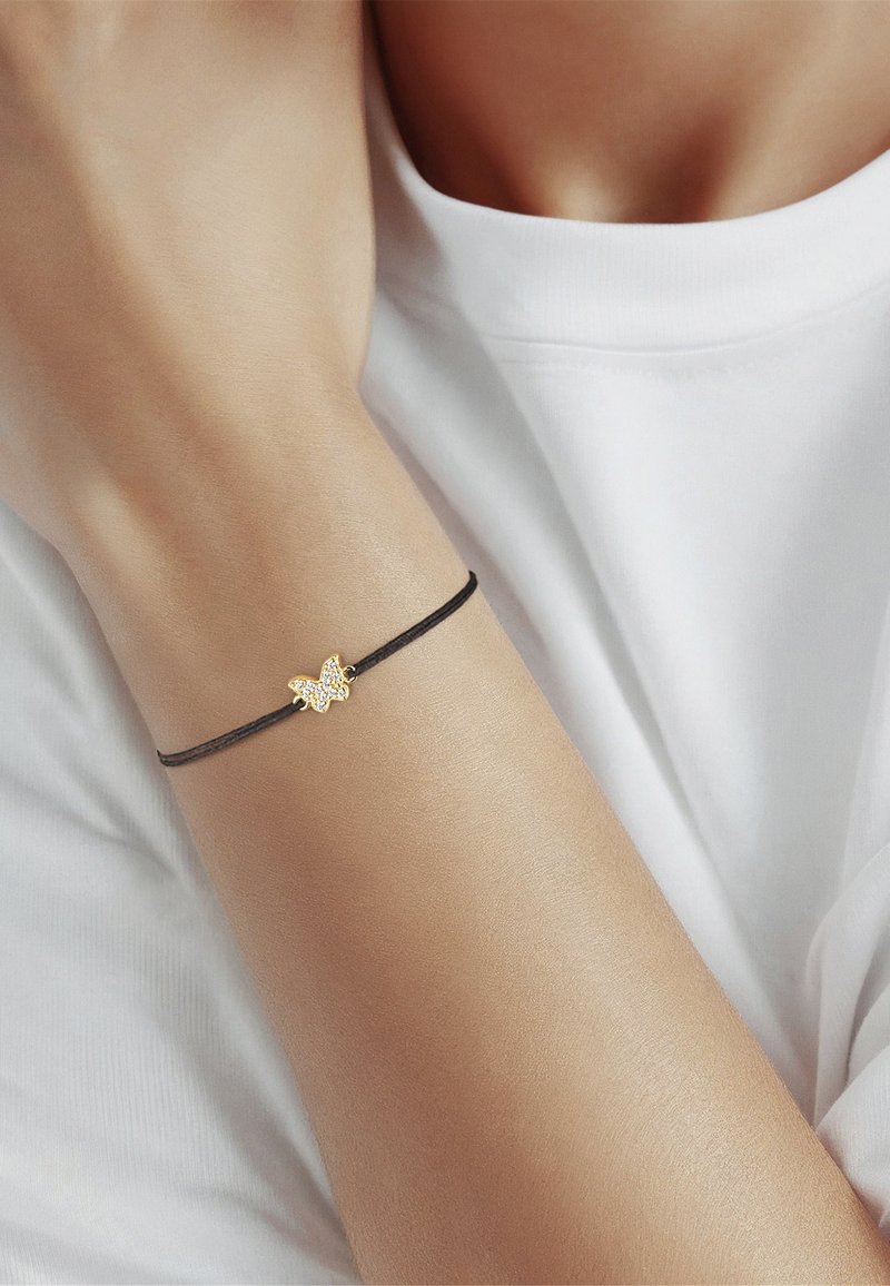 Melna aukliņu aproce ar zelta tauriņu piekariņu, kas rotāts ar caurspīdīgām akcentiem, uz gludas, gaišas ādas rokas pret baltu auduma fona.