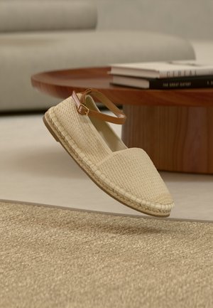 Espadrille beige tissée avec bride marron à la cheville flottant au-dessus d'un tapis beige texturé, avec une table basse en bois et des livres en arrière-plan.