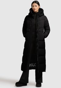Dlhý čierny puffový kabát s kapucňou, s prešívanými časťami, predným zipsom a bočnými vreckami. Nosený cez zodpovedajúci outfit.