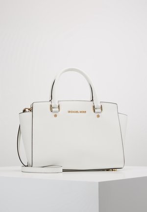 Weiße Michael Kors Handtasche mit goldenen Beschlägen, doppelten Griffen und abnehmbarem Schulterriemen auf weißem Podest vor einfachem Hintergrund.