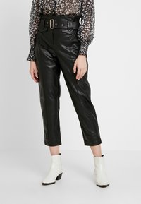 Svart läderbyxor med midja av bälte, kort längd och framfickor, kombinerat med en transparent mönstrad blus och vita ankelstövlar.