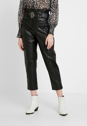 Zwarte leren broek met een ceintuur in de taille, cropped lengte en voorzakken, gecombineerd met een doorschijnende blouse met patroon en witte enkellaarsjes.