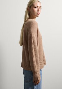 Junge Frau mit langem, blonde Haar trägt einen braunen, flauschigen Pullover und blaue Jeans, steht seitlich gegen eine weiße Wand.