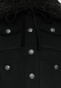 Fekete gyapjúkabát textúrált fekete gallérral, ezüst koponya gombokkal és húzózsinórral a nyaknál. Két elülső zsebbel rendelkezik.