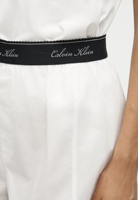 Μαύρη ελαστική ζώνη Calvin Klein σε λευκό ένδυμα φορεμένο στη μέση με εμφανές χέρι στο πλάι