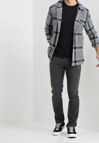 Chemise à carreaux noir et blanc, t-shirt à col rond noir, jeans gris et baskets noires avec semelles blanches. Look décontracté et design superposé.