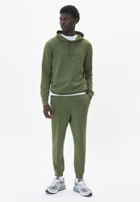 Sudadera de color verde oliva con cordones, combinada con pantalones ajustados a juego. Llevada con zapatillas deportivas grises que tienen acentos en blanco.