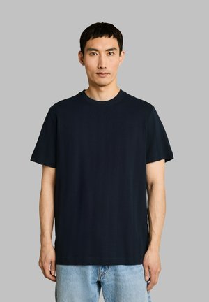 Jonge man met kort zwart haar, die een effen zwart T-shirt met ronde hals en lichtblauwe spijkerbroek draagt tegen een grijze achtergrond.