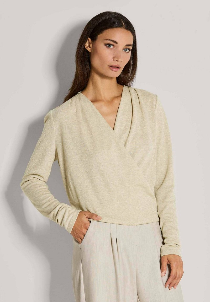 Helle beige Langarm-Wickeloberteil mit tiefem V-Ausschnitt, weichem Material und drapiertem Vorderteil, kombiniert mit hellen beigen, plissierten Hosen.