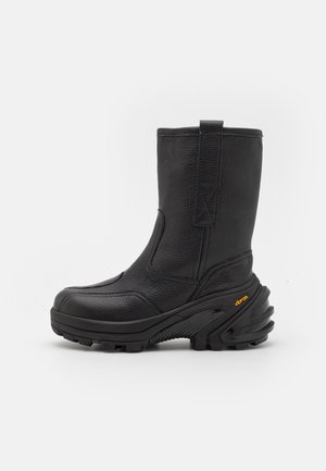 Crna kožna gležnjača s teksturiranim završetkom, zakrivljenim dizajnom potplata, ojačanim vrhom i istaknutim Vibram logom sa strane.