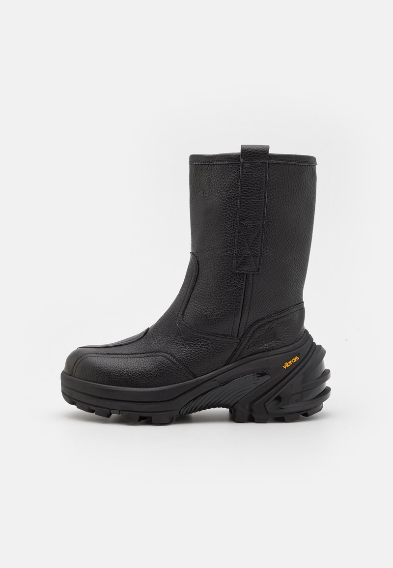 Bota de tobillo de cuero negro con un acabado texturizado, diseño de suela curva, puntera reforzada y un prominente logo de Vibram en el lateral.