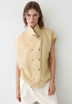 Frau mit hellbraunen Haaren trägt eine ärmellose beige Weste mit hohem Kragen und Metallknöpfen, kombiniert mit lockeren weißen Hosen, stehend an einer weißen Wand.