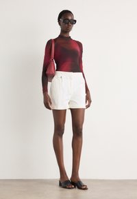 Red and black sheer long-sleeve top, white tailored shorts with front button, small red handbag, and black open-toe slide sandals.
