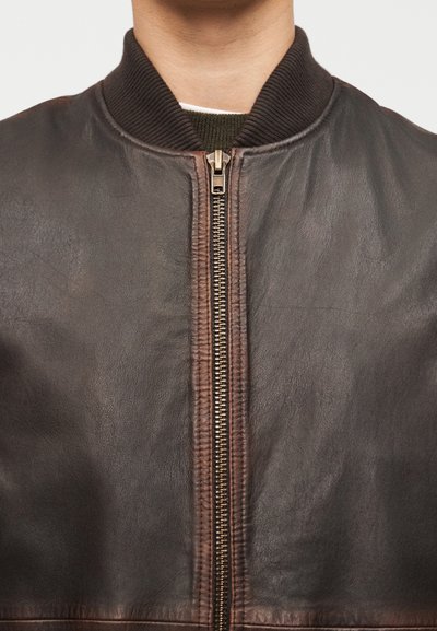 Κοντινό πλάνο ενός ατόμου που φοράει σκούρο καφέ δερμάτινο μπουφάν με χρυσαφί φερμουάρ και ριμπ γιακά πάνω από ένα πράσινο πουλόβερ.