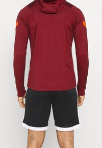 Bordeauxfarbenes Langarm-Hoodie mit Kapuze und orangefarbenem Akzent auf der Schulter, kombiniert mit schwarzen Shorts mit weißem Seitenbesatz und strukturiertem Material.