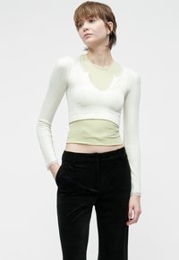 Młoda osoba z krótkimi włosami, ubrana w biały krótki kardigan na jasnej zielonej bluzce oraz czarnych spodniach, stoi z jedną ręką w kieszeni.