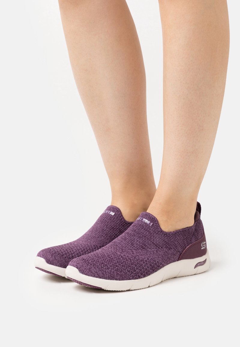 Jambes portant des baskets tricotées violettes à enfiler avec des semelles blanches sur fond blanc, montrant les vues latérale et frontale.