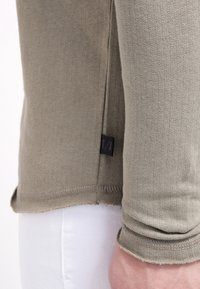 Olijfgroene langemouwenshirt, geribbelde textuur, zijsplitdetails en een klein zwart label aan de zoom. De stof lijkt zacht en rekbaar.