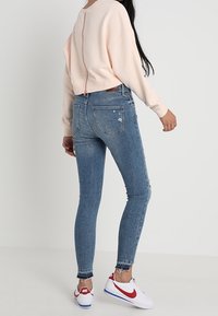 Felpa corta rosa chiaro con cerniera sul retro, abbinata a jeans skinny blu con dettagli strappati e orli sfilacciati; sneaker bianche.