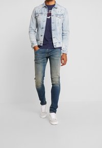 Light blue denim jacket with distressed details, navy t-shirt, faded blue jeans, and white sneakers. Casual, fitted style.