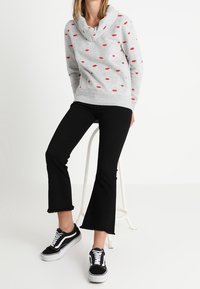 Grauer Kapuzenpullover mit roten Lippenmustern und Schlauchkragen. Dazu schwarze gekürzte Jeans und schwarze Sneakers mit weißen Sohlen.