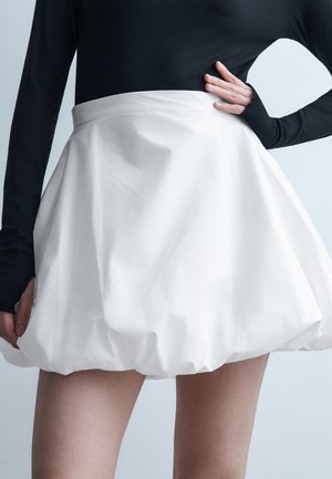 Woman wearing a white, voluminous bubble skirt and a fitted long-sleeve black top, posing with one hand on her hip.