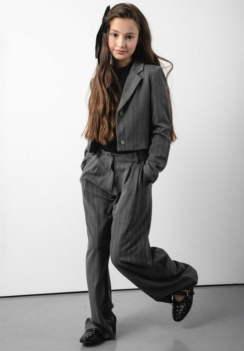 Szary garnitur w prążki z dopasowaną marynarką i luźnymi spodniami, połączony z czarnym topem i czarnymi butami z ćwiekami. Długie włosy z czarną kokardą.