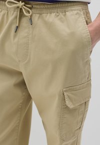 Pantalones cargo caqui hechos de una tela ligera; cuentan con una cinturilla elástica con cordón, bolsillos laterales y un gran bolsillo cargo.