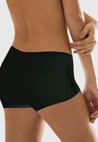 Rückansicht einer Person, die schwarze nahtlose Boyshort-Unterwäsche trägt, während eine Hand sanft den gegenüberliegenden Arm berührt, vor einem schlichten Hintergrund.