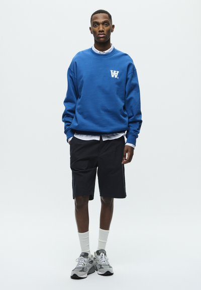Sudadera azul con logo blanco, pantalones cortos negros, camisa blanca de botones debajo, zapatos grises y calcetines blancos. Diseño atlético simple.