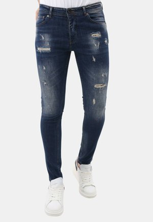 Donkerblauwe slim-fit jeans met gescheurde en vervaagde details, gecombineerd met witte sneakers en een wit shirt.