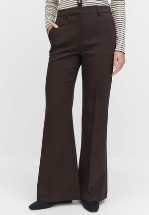 Vrouw in donkerbruine wijde broek, gestreept lange mouwen shirt, zwarte schoenen, staand met de linkerhand in de zak tegen een effen achtergrond.