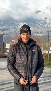 Juoda pūkinė striukė su siuvinėta tekstūra, dėvima ant juodo megztinio su užtrauktuku. Juodas vilnonis kepurė su baltu raštu. Neutralus lauko fonas.