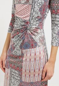 Musterkleid mit mehrfarbigen Designs in Rot, Blau und Schwarz, mit einem gedrehten Knoten-Detail in der Taille und Dreiviertelärmeln.