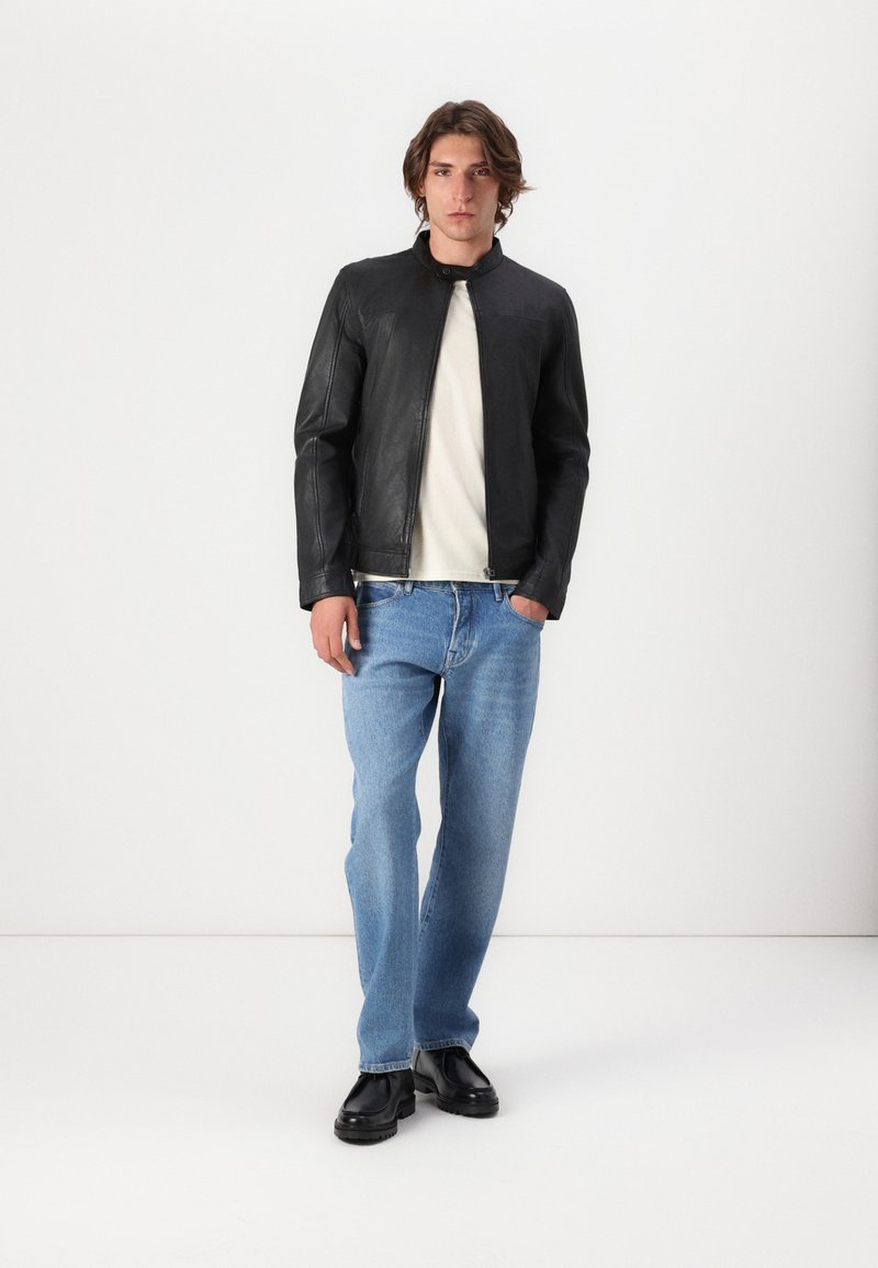 Schwarze Lederjacke mit Stehkragen, kombiniert mit hellblauen gerade geschnittenen Jeans und schwarzen Schnürschuhen vor einem neutralen Hintergrund.