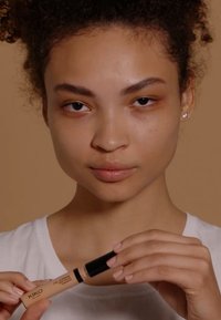 Correcteur couvrant KIKO dans un tube beige avec un bouchon noir, tenu par une personne aux cheveux bouclés portant une chemise blanche, sur un fond beige.