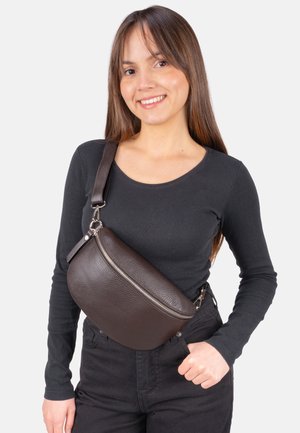 Jeune femme aux longs cheveux bruns portant un t-shirt noir à manches longues et un grand sac bandoulière en cuir marron foncé avec une fermeture éclair.