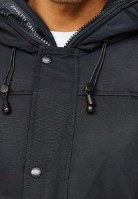 Chaqueta negra con capucha que presenta un cierre de cremallera y snaps, cordones y una textura suave, con un sutil detalle del logo en el cuello interior.