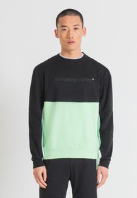Sudadera color bloqueada en negro y verde menta con cuello redondo, mangas largas y un pequeño logo impreso en la parte frontal. Textura de tela suave.