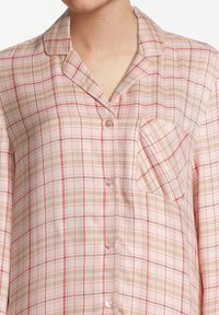 Vrouw in een roze, beige en rode geblokte blouse met een klein borstzakje en een getailleerde kraag.