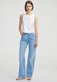 Witte mouwloze top met kantdetails, gecombineerd met lichtblauwe flared jeans met een stippenpatroon en subtiele vervaging.