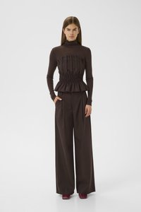 Giovane donna che indossa un top peplum plissettato marrone scuro, pantaloni a gamba larga e scarpe bordeaux, in piedi con una mano in tasca.