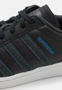 Svart lädersneaker med grå tygremsor, blå sömmar och "SUPERSTAR"-logotyp. Har runda snören och vit gummisula.