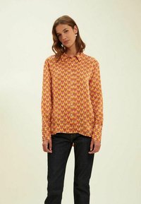 Chemise à manches longues à motifs floraux orange et jaune, avec un boutonnage à l'avant, en tissu doux, col classique et coupe droite. Portée avec un jean foncé.