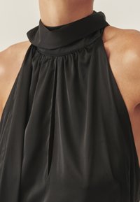 Top negro de cuello halter con cuello alto, presenta un frente plisado y una tela lisa y ligera. Sin herrajes visibles ni patrones.